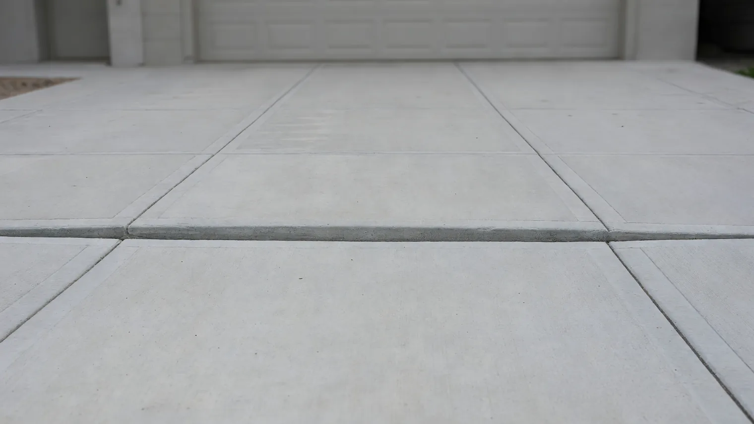 Concrete flatwork meeting a driveway apron at a stepped joint, the near slab sitting below the fixed edge, a white garage door out of focus behind.